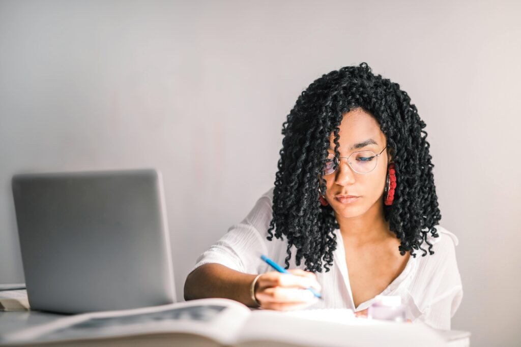 Focused young woman working at a desk with a laptop and writing notes.